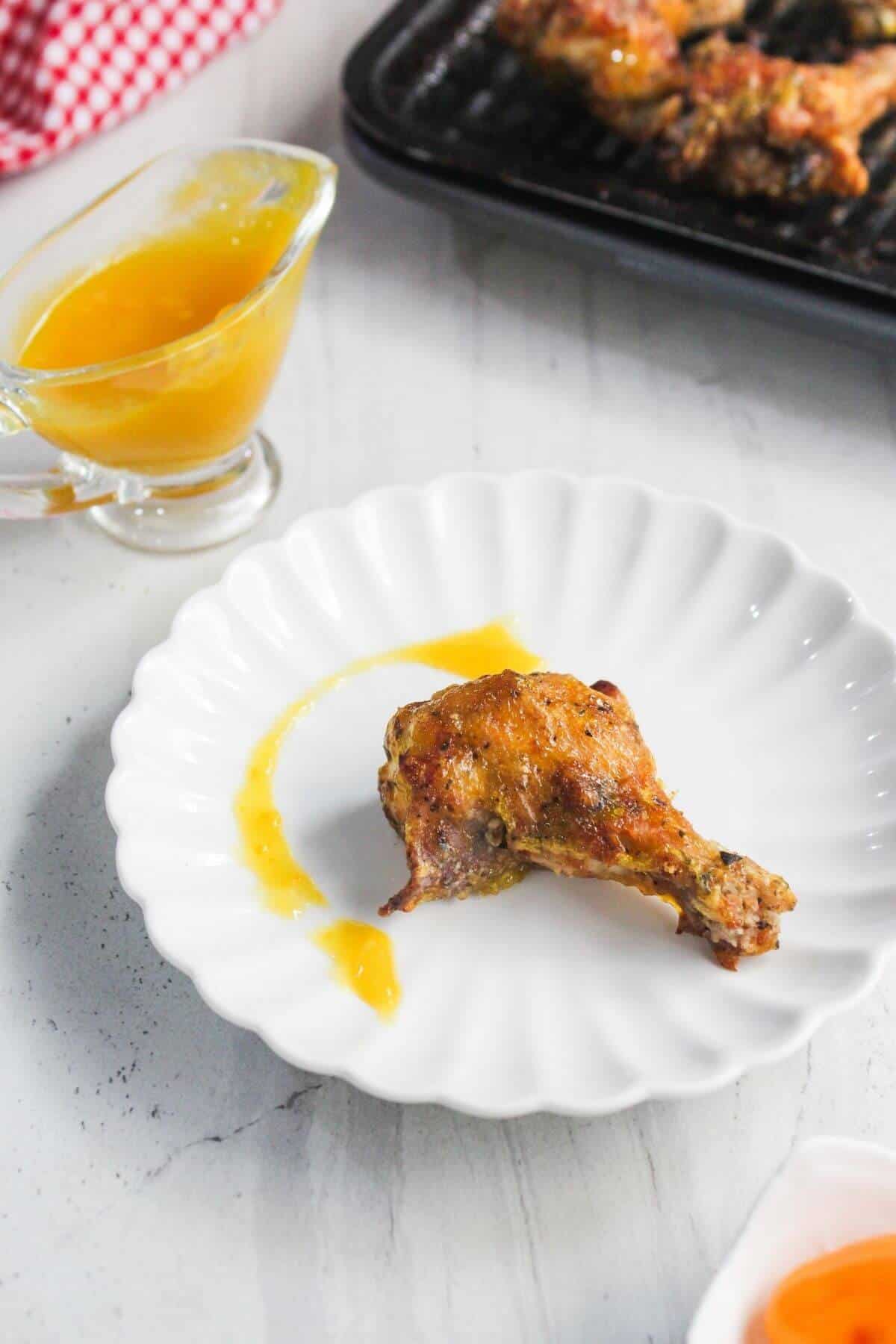 A cooked chicken drumstick with sauce sits on a white plate, with extra sauce in a small glass pitcher nearby. Grilled chicken pieces are visible in the background.