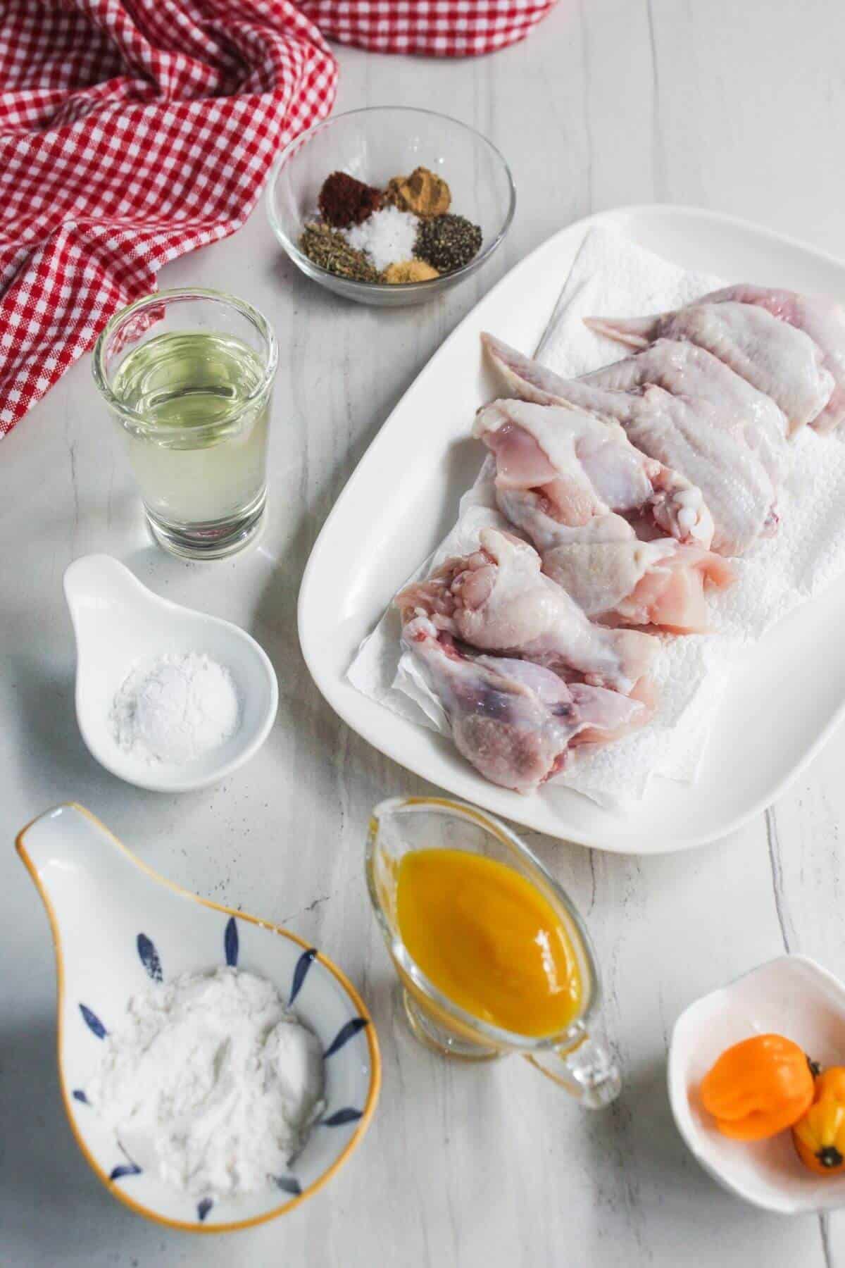 Raw chicken wings on a plate with paper towels, surrounded by small bowls of flour, spices, oil, yellow sauce, and a red-checkered cloth on a white surface.