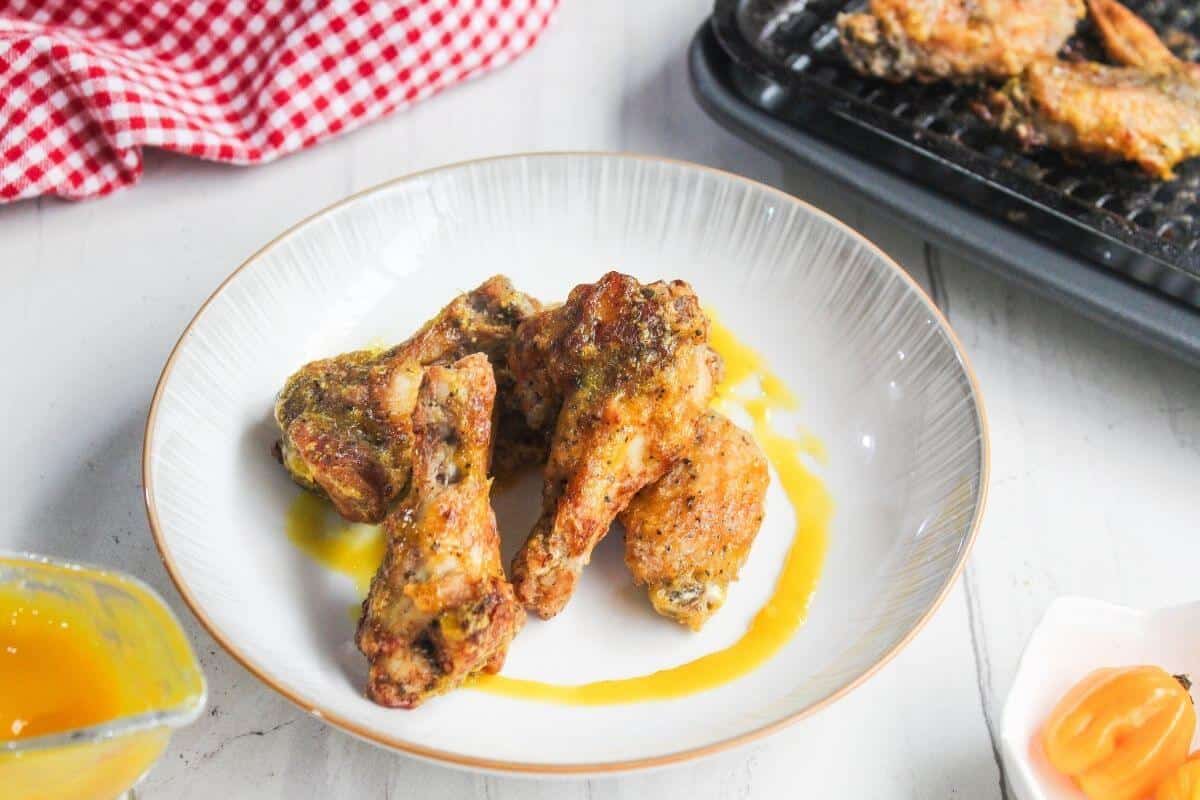A white plate with several cooked chicken wings drizzled with yellow sauce, placed on a white table next to a red checkered cloth and a tray of more wings.