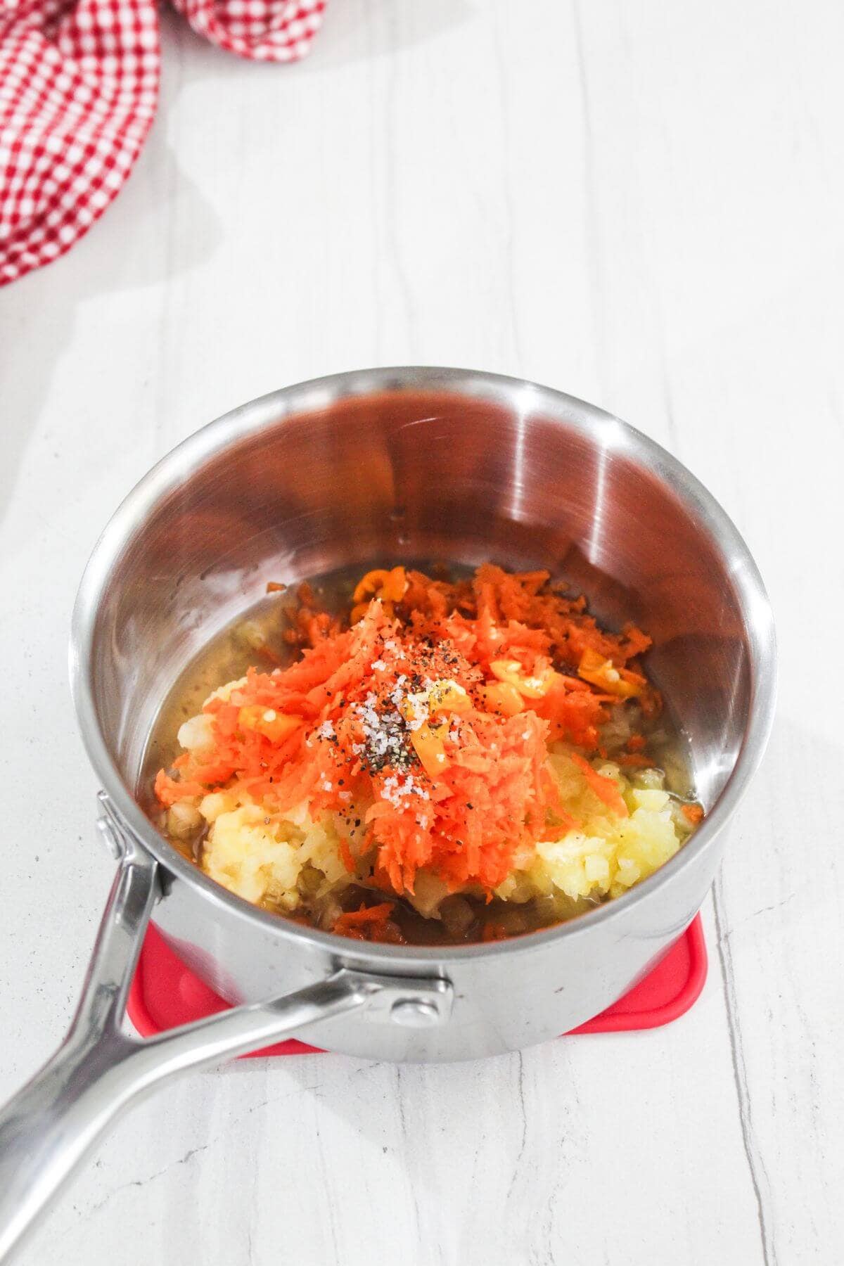 A stainless steel saucepan with grated carrots, mashed potatoes, and seasoning on a white countertop.