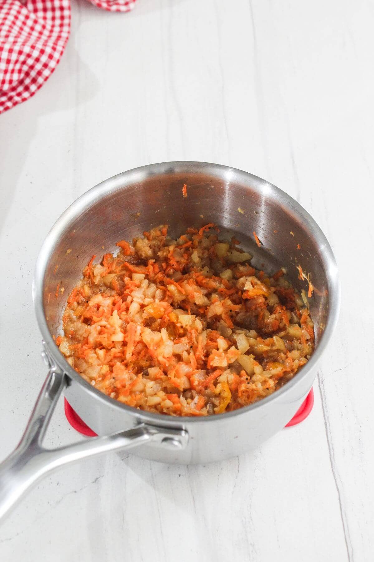A stainless steel saucepan containing sautéed chopped onions and carrots sits on a white countertop.