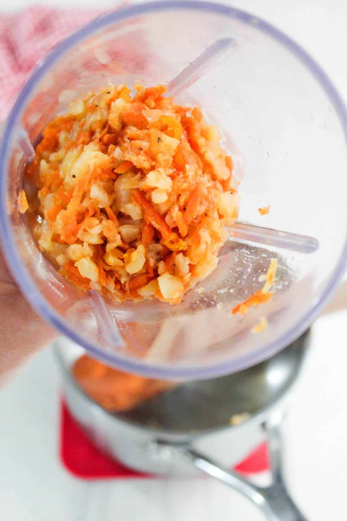 Close-up of a food processor cup with chopped onions and carrots inside, held above a pot on a kitchen countertop.