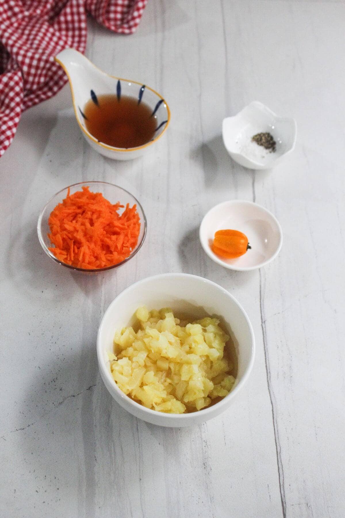 Chopped pineapple, grated carrot, a whole habanero pepper, vinegar, pepper, and salt are arranged in small bowls on a white surface.