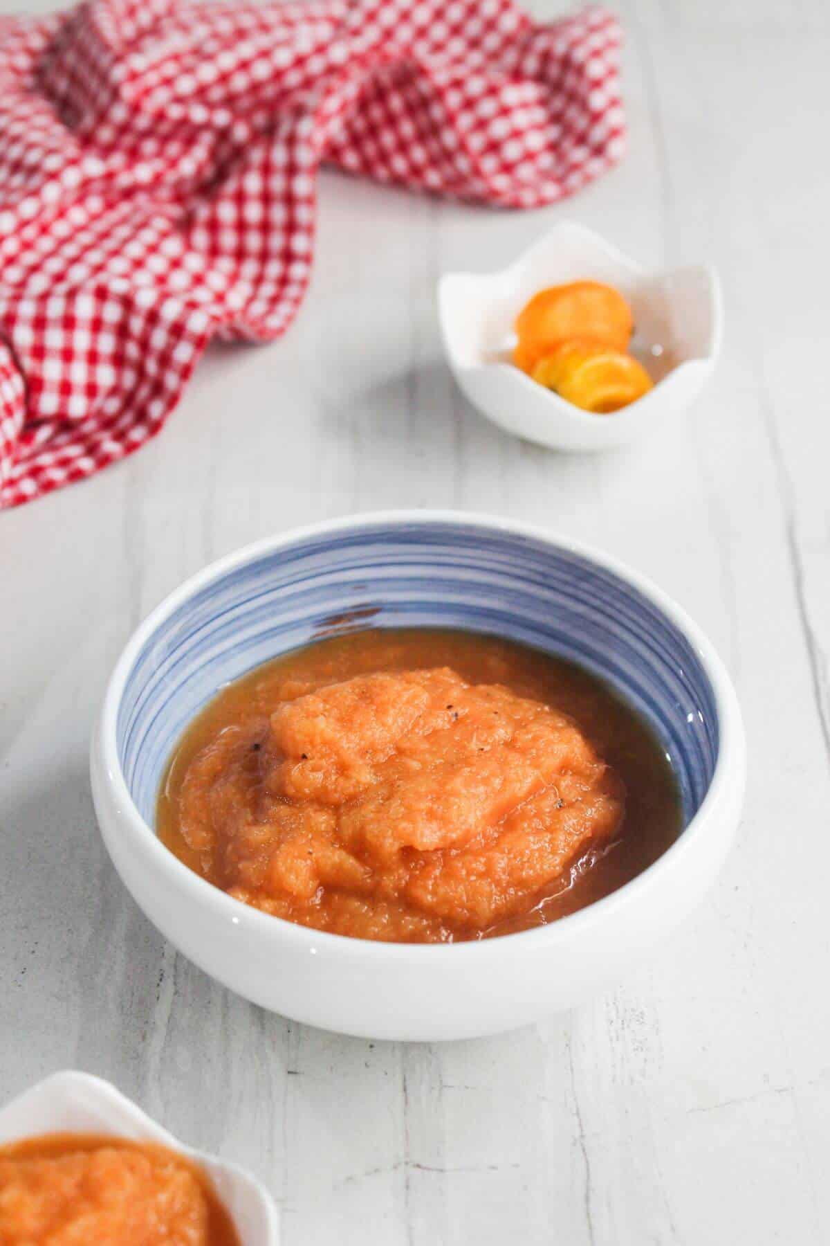 A bowl of orange fruit puree sits on a light surface, with a small dish of sliced fruit and a red checkered cloth in the background.