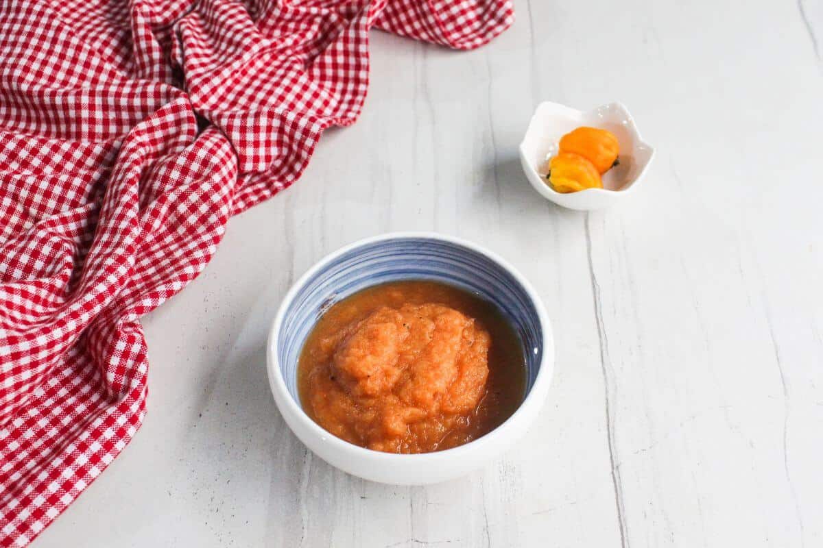 A bowl of orange sauce sits on a light surface beside a small dish holding two yellow peppers and a red checkered cloth.