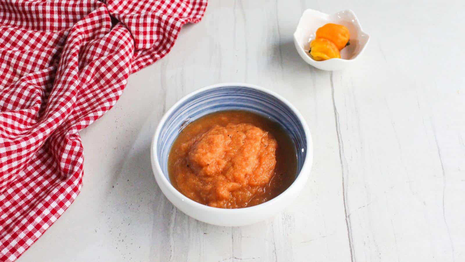 A bowl of orange pepper sauce sits on a white surface next to a small dish with two whole yellow peppers and a red checkered cloth.