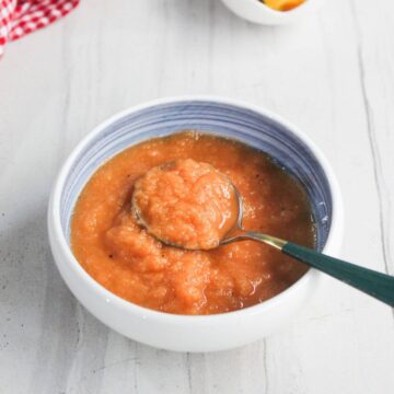 A bowl of thick, orange-colored applesauce with a spoon resting inside, placed on a light-colored surface.