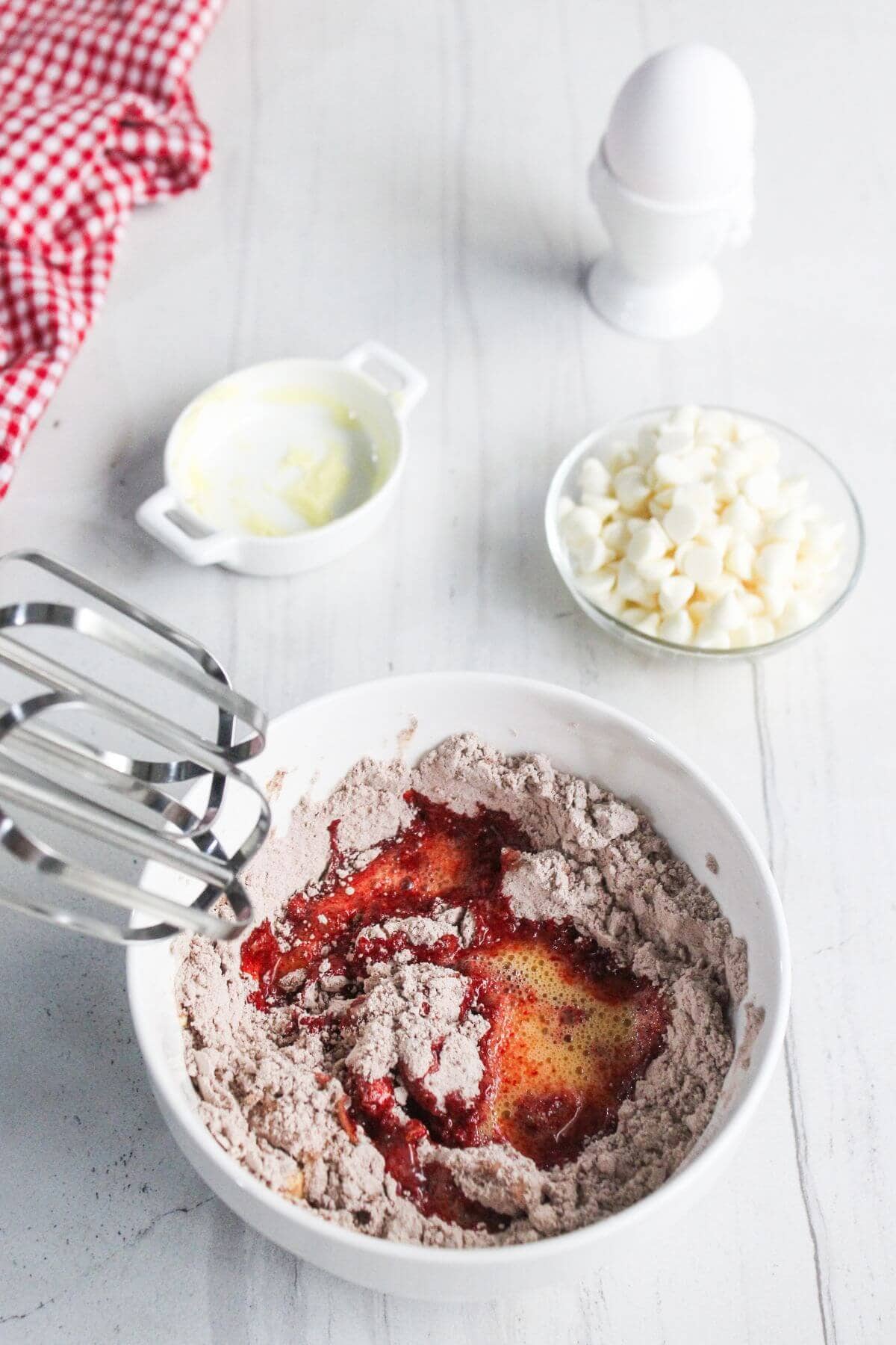 A mixing bowl with cake mix, eggs, and red liquid, an electric mixer, a bowl of white chocolate chips, an egg in a holder, and a butter dish on a white surface.