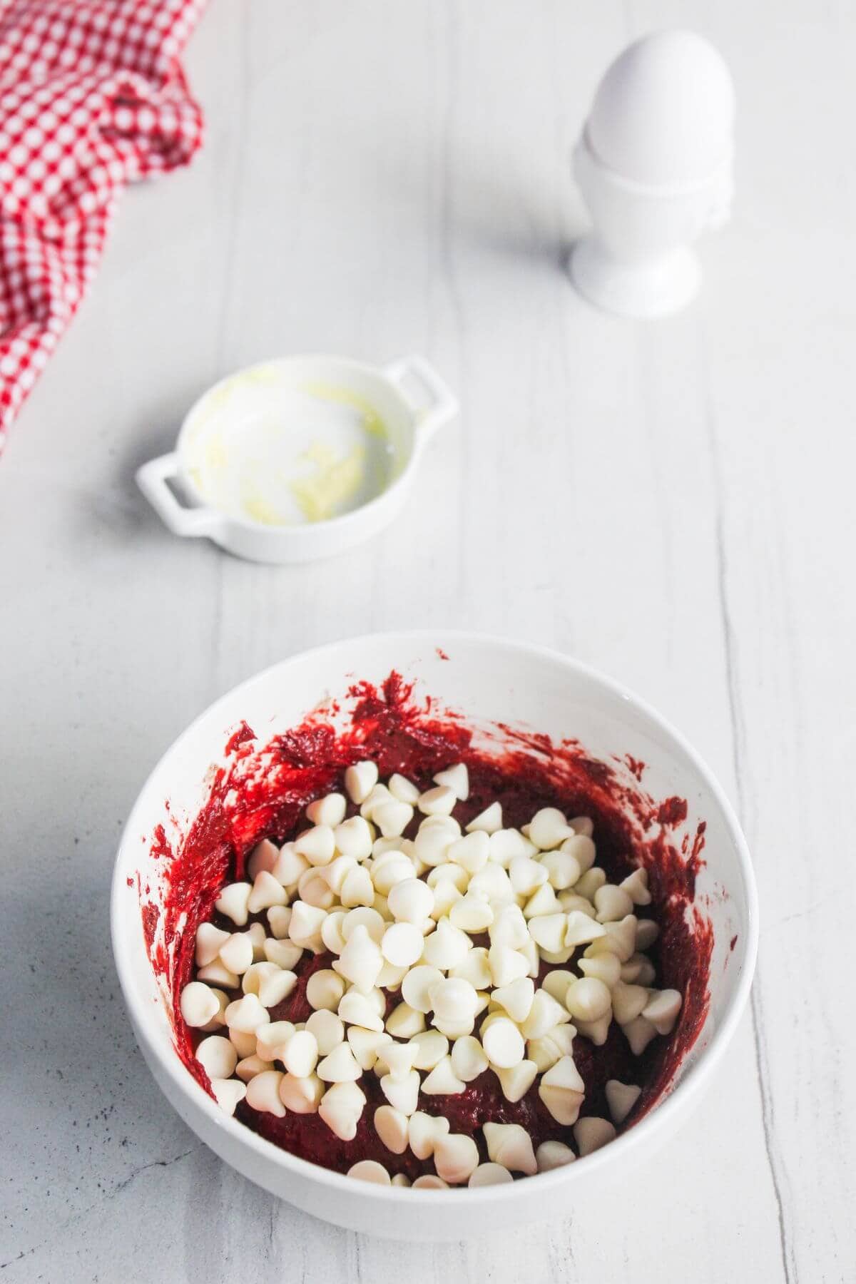 A white bowl filled with red batter and white chocolate chips sits on a light countertop. A dish with butter, an egg in an egg cup, and a red checkered cloth are in the background.