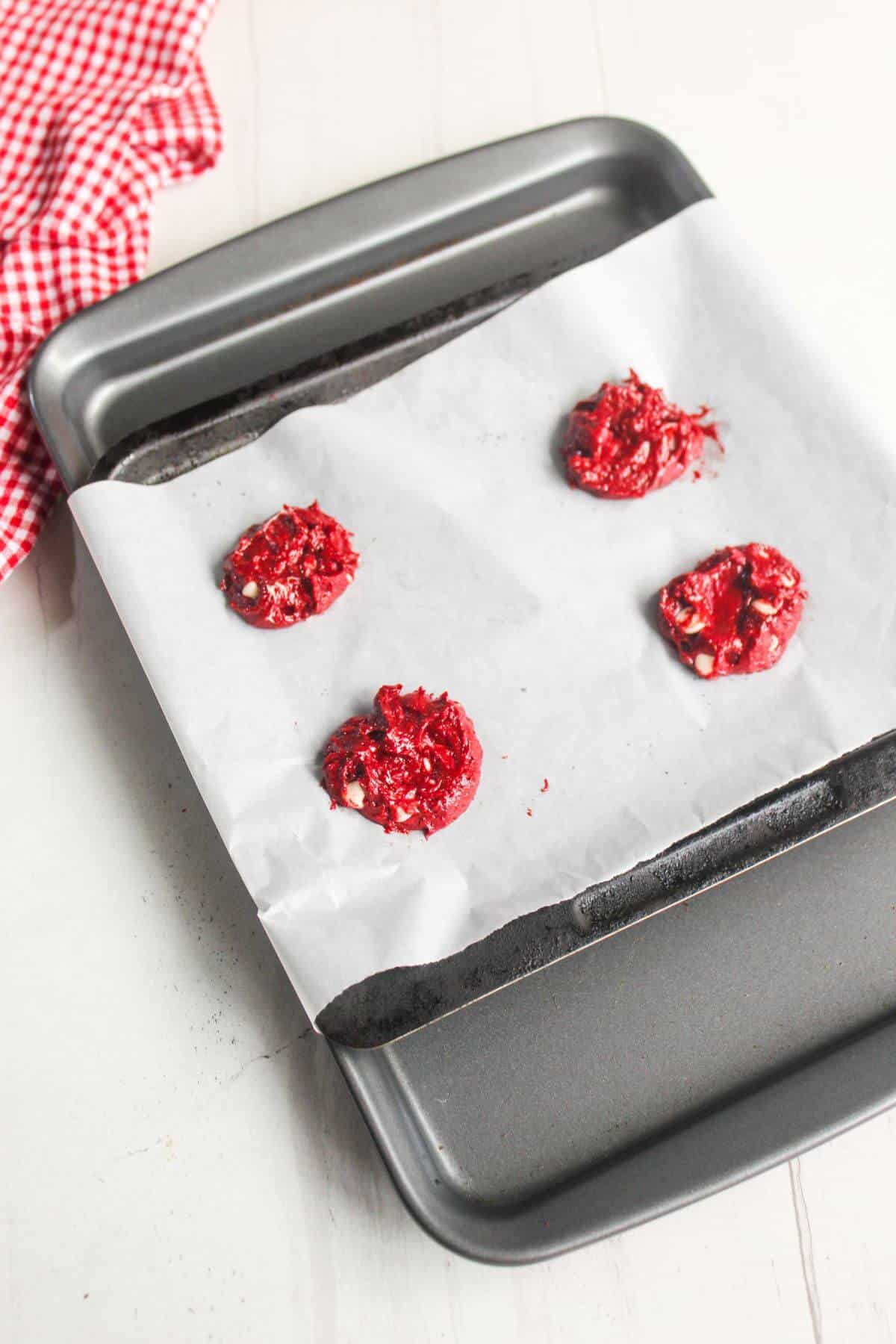 A baking tray lined with parchment paper holds four scoops of red cookie dough, ready to be baked. A red checkered cloth is in the upper left corner.