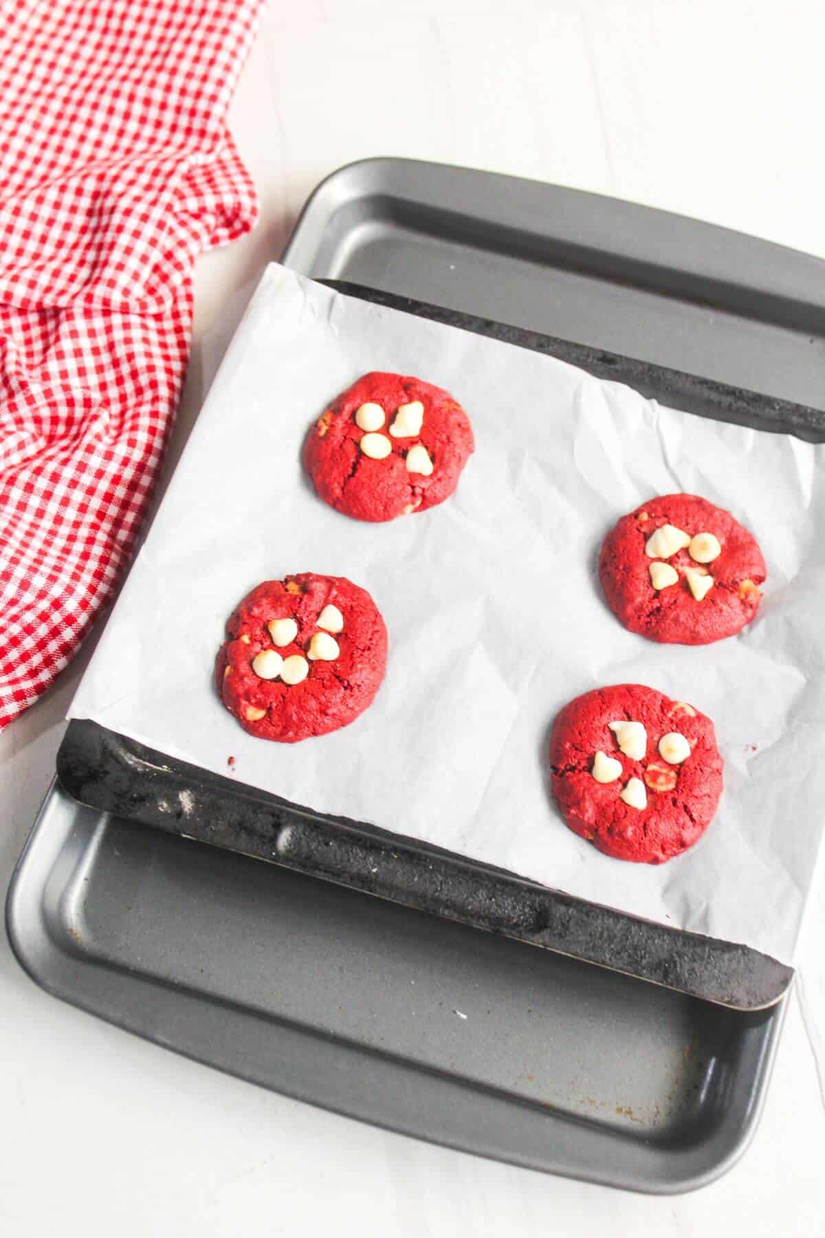 A baking tray with four red cookies topped with white chocolate chips on parchment paper, next to a red and white checkered cloth.