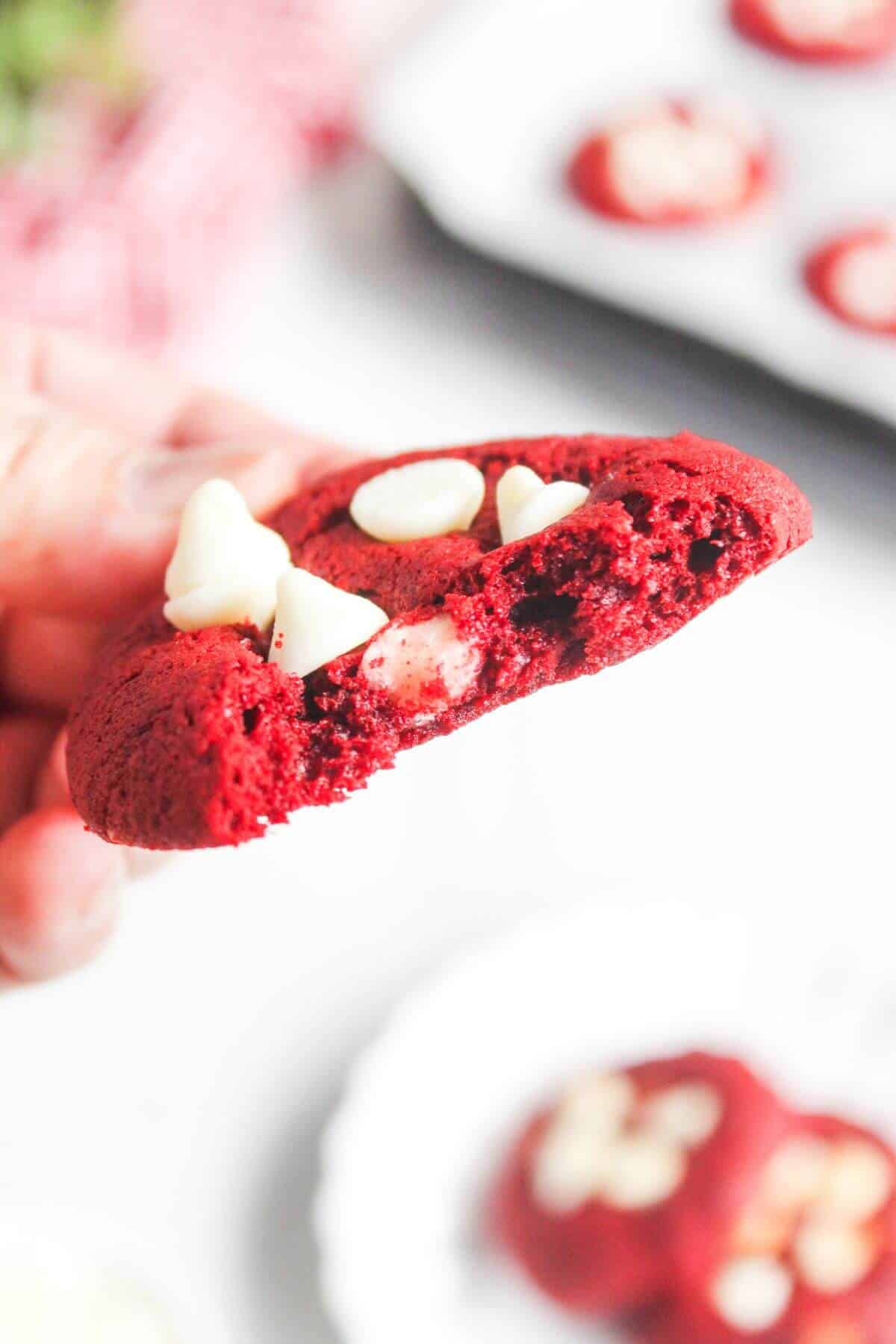 A hand holds a bitten red velvet cookie with white chocolate chips. Other similar cookies are visible in the background on a plate and tray.