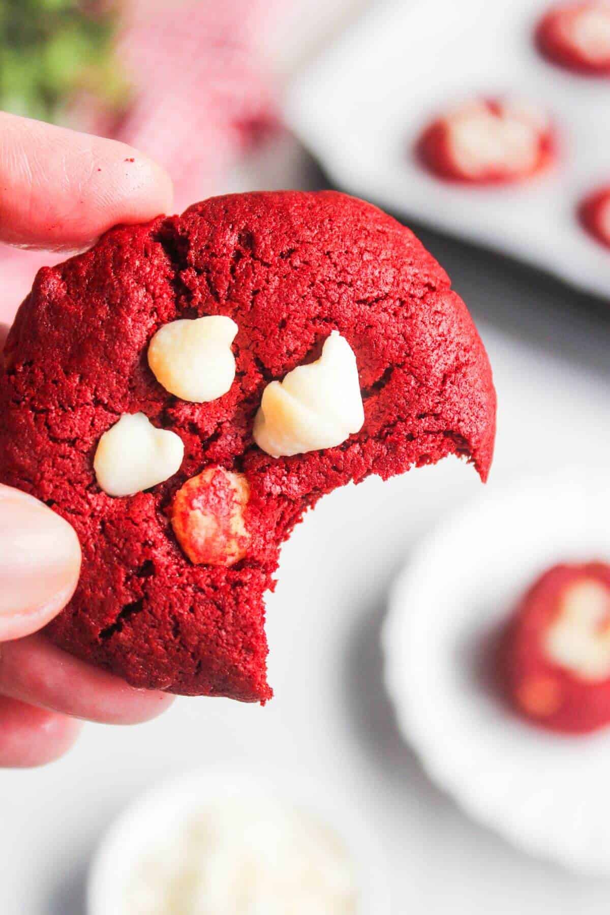 A hand holds a red velvet cookie with white chocolate chips and a bite taken out of it. Other cookies and white chocolate chips are visible in the background.