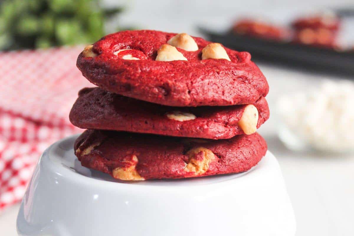 Three red velvet cookies with white chocolate chips stacked on a white dish, with a red checkered cloth and blurred background.