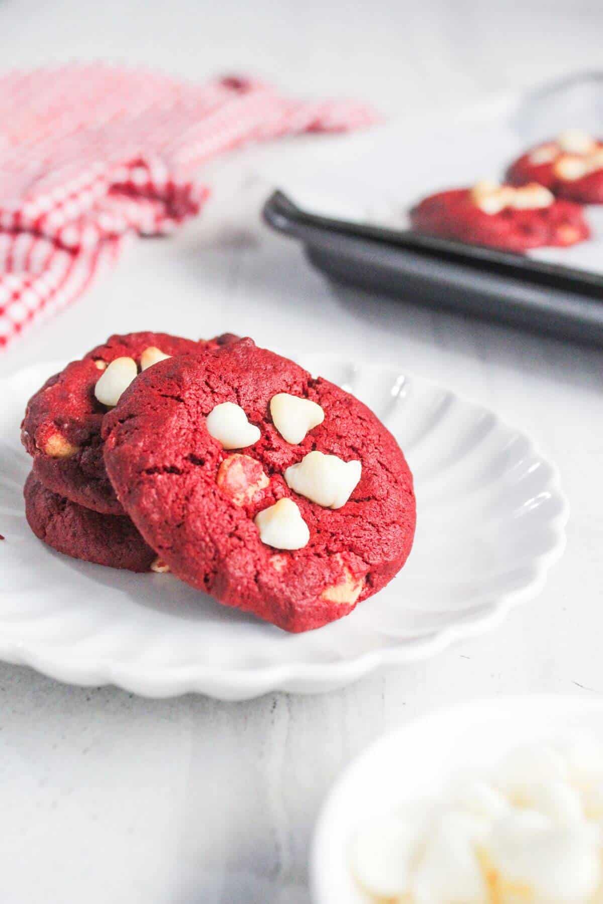 Three red velvet cookies with white chocolate chips on a white scalloped plate, with a baking tray and a red checked cloth in the background.