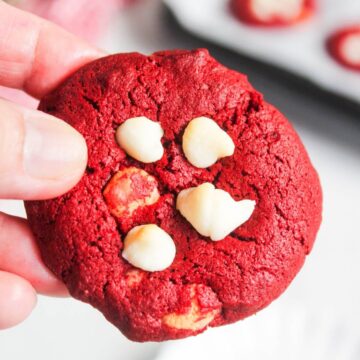 A hand holding a red velvet cookie with white chocolate chips; more cookies are blurred in the background.