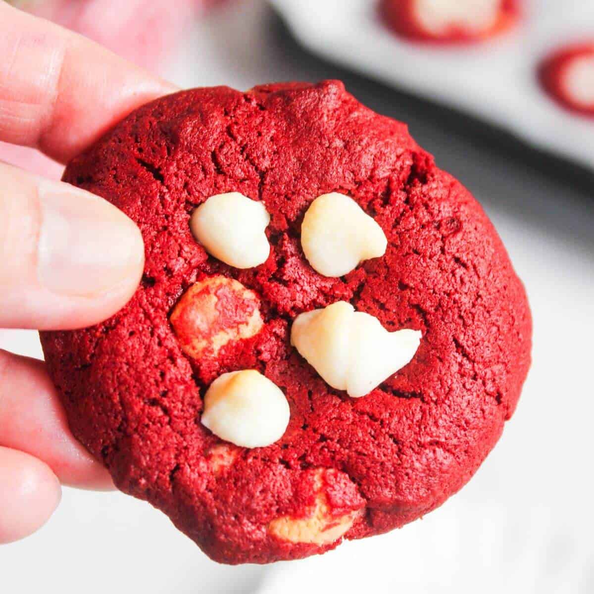 A hand holding a red velvet cookie with white chocolate chips; more cookies are blurred in the background.
