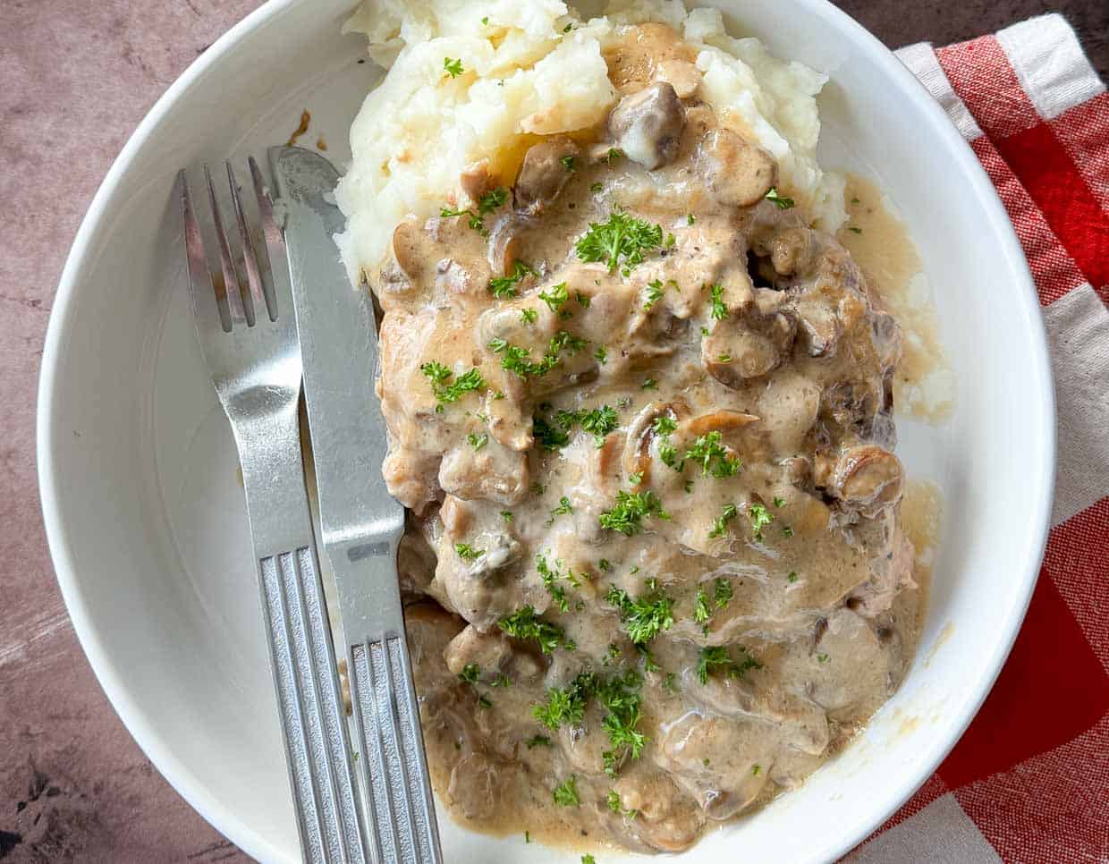A plate of mashed potatoes topped with beef and creamy mushroom gravy, garnished with chopped parsley, with a fork and knife on the side.