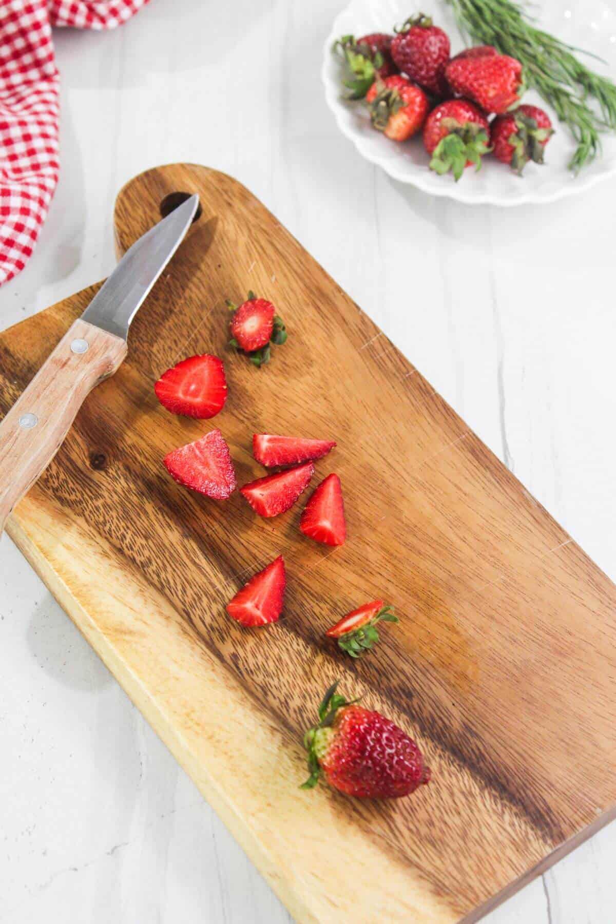 A wooden cutting board with sliced strawberries and a paring knife, next to a plate of whole strawberries on a white countertop.