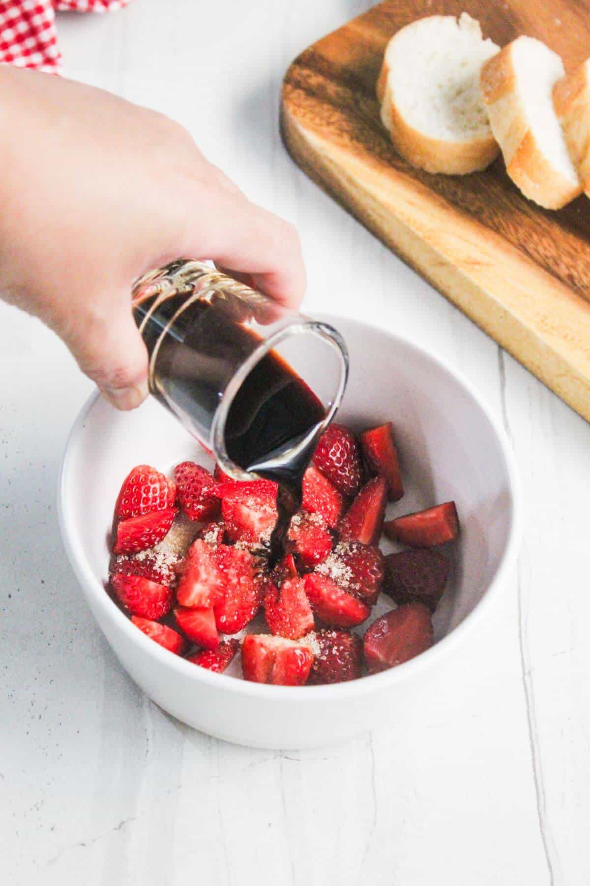 A hand pours balsamic vinegar over sliced strawberries in a white bowl, with sliced bread on a wooden board in the background.