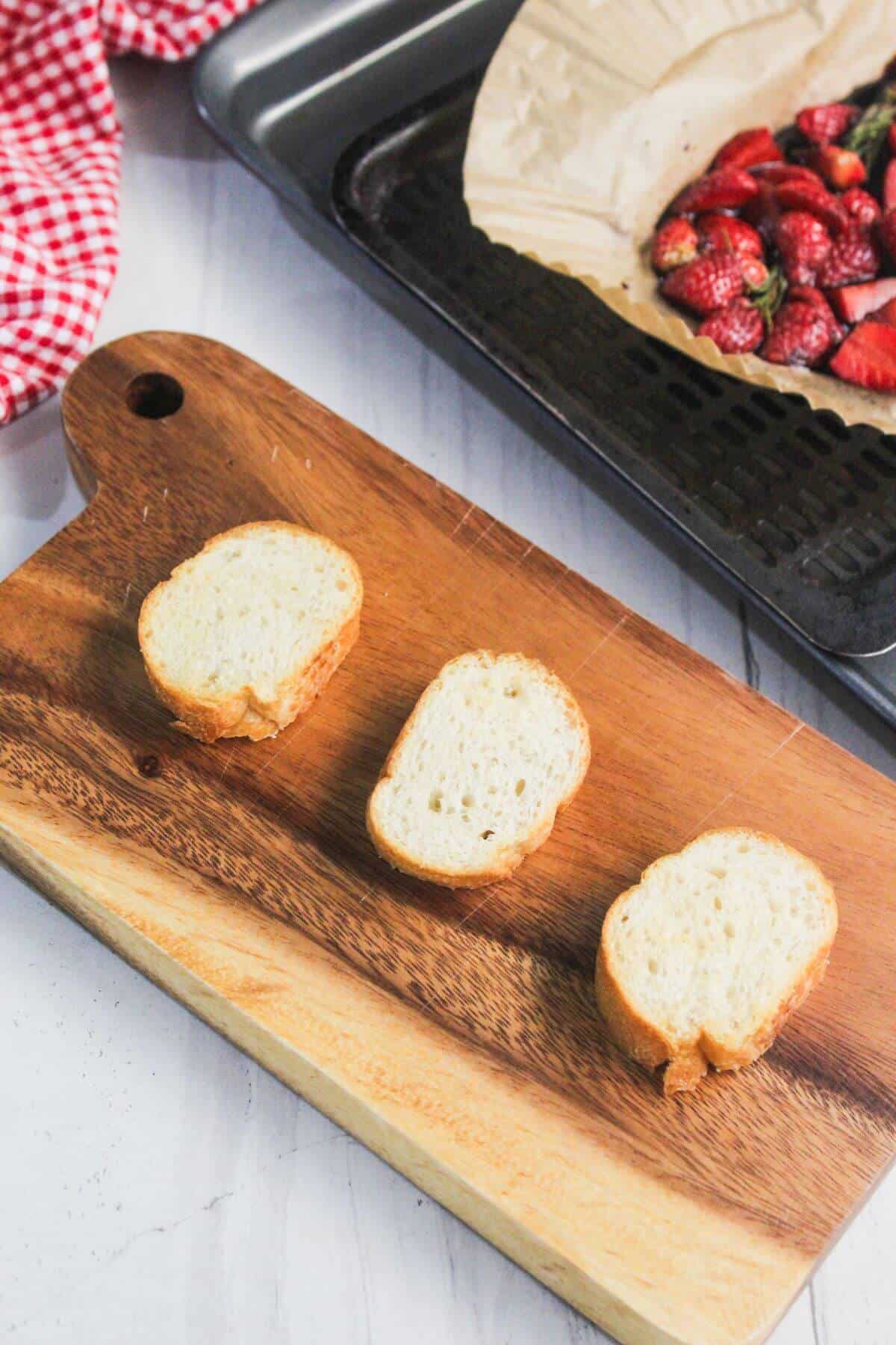 Three slices of baguette on a wooden cutting board with a baking tray of roasted strawberries in the background.