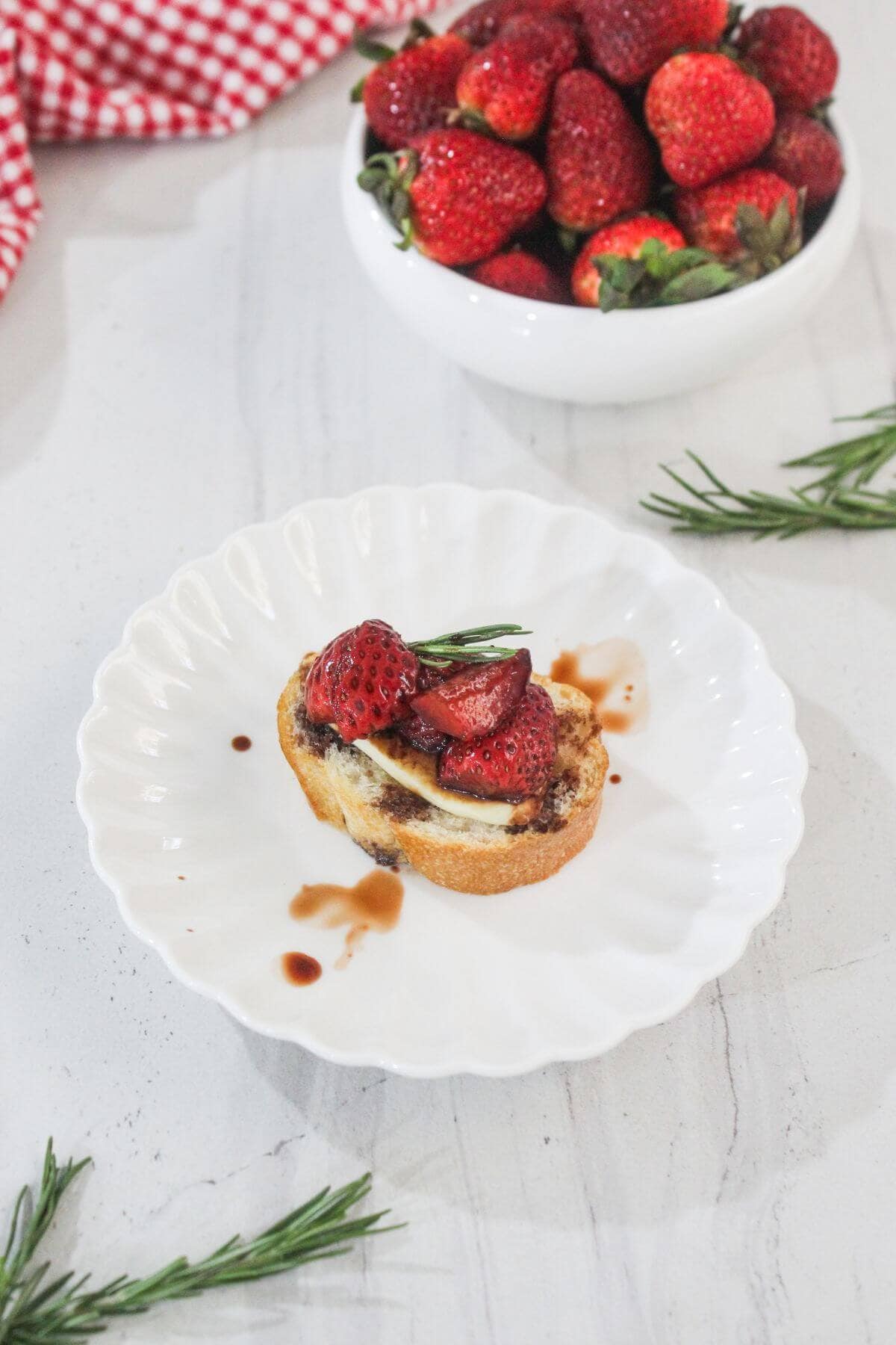 A slice of bread topped with cheese, strawberries, and herbs is drizzled with sauce on a white plate; a bowl of fresh strawberries and sprigs of rosemary are nearby.
