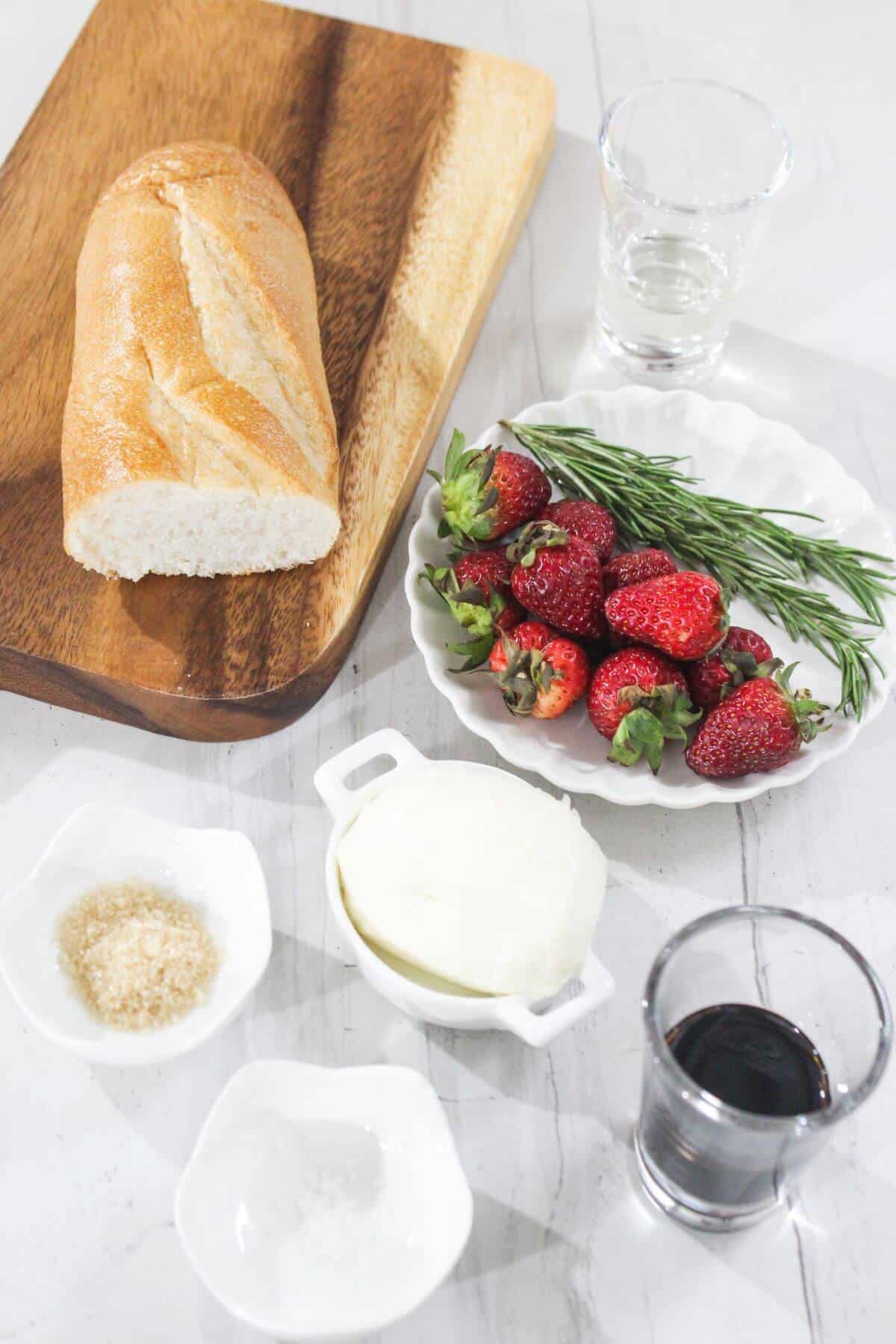 A loaf of bread on a wooden board, strawberries, rosemary, mozzarella, balsamic vinegar, sugar, and salt arranged on a white surface.