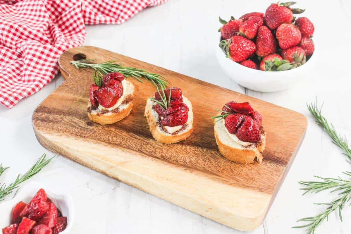 Three pieces of toasted bread topped with cream, strawberries, and rosemary are arranged on a wooden board. A bowl of strawberries and a red checkered cloth are nearby.