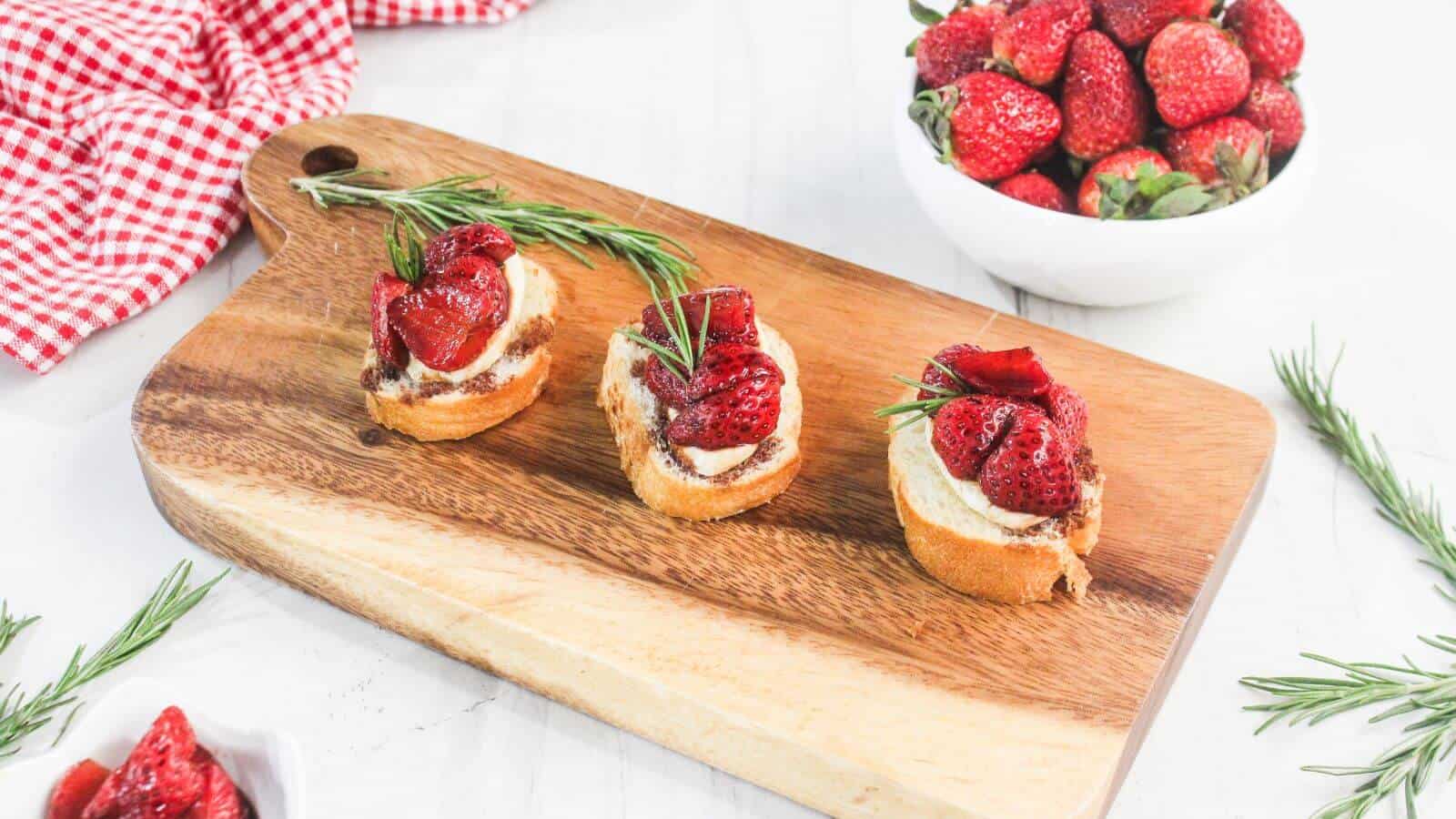 Three pieces of toasted bread topped with cream, strawberries, and rosemary are arranged on a wooden cutting board, with a bowl of strawberries and a red-checkered cloth nearby.
