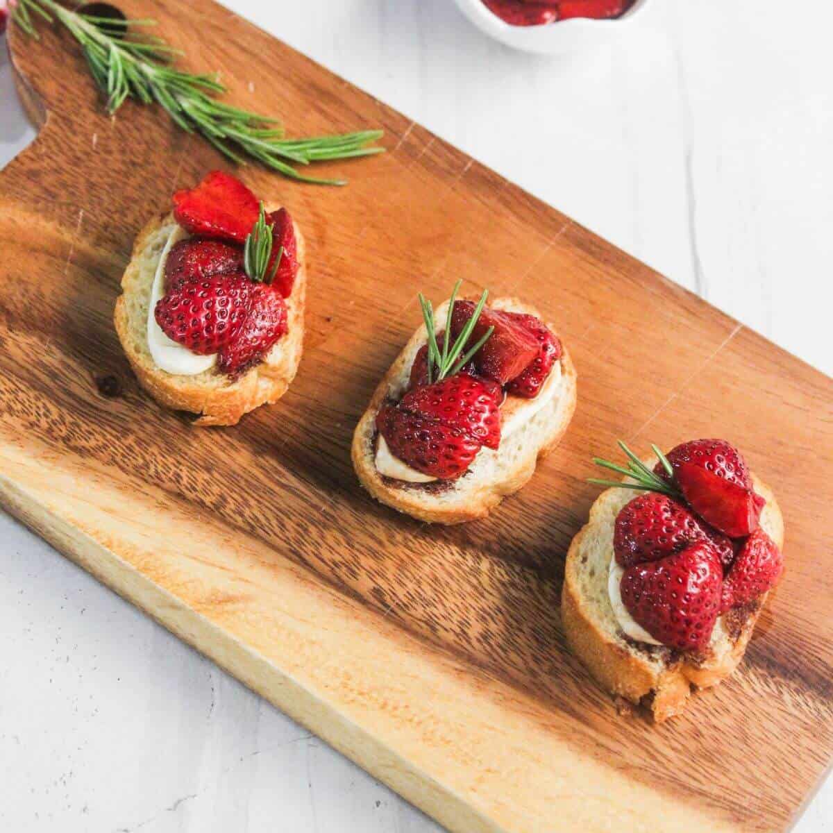 Three pieces of toasted bread topped with cream, strawberries, and rosemary are arranged in a row on a wooden serving board.