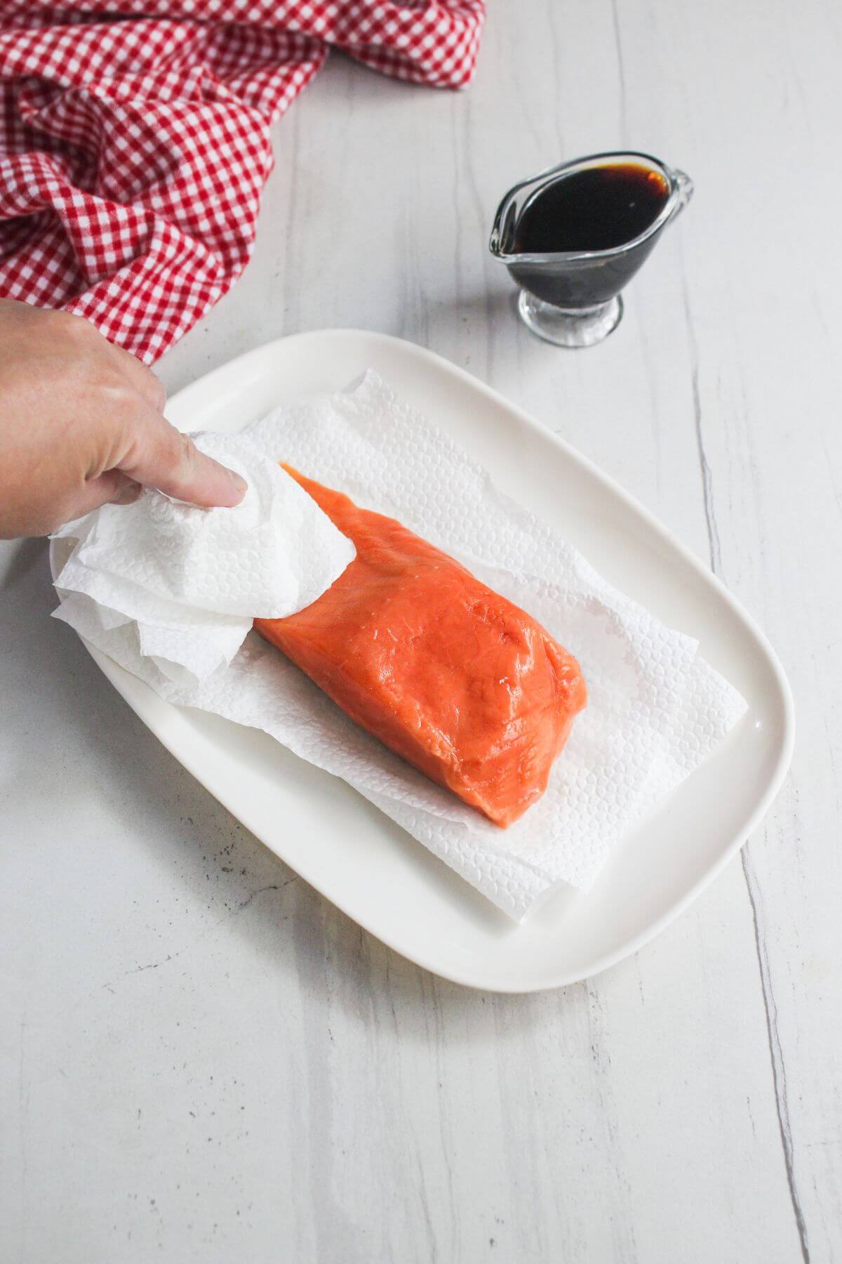 A hand patting a raw salmon fillet dry with paper towels on a white plate, with a small glass container of dark sauce and a red checkered cloth nearby.