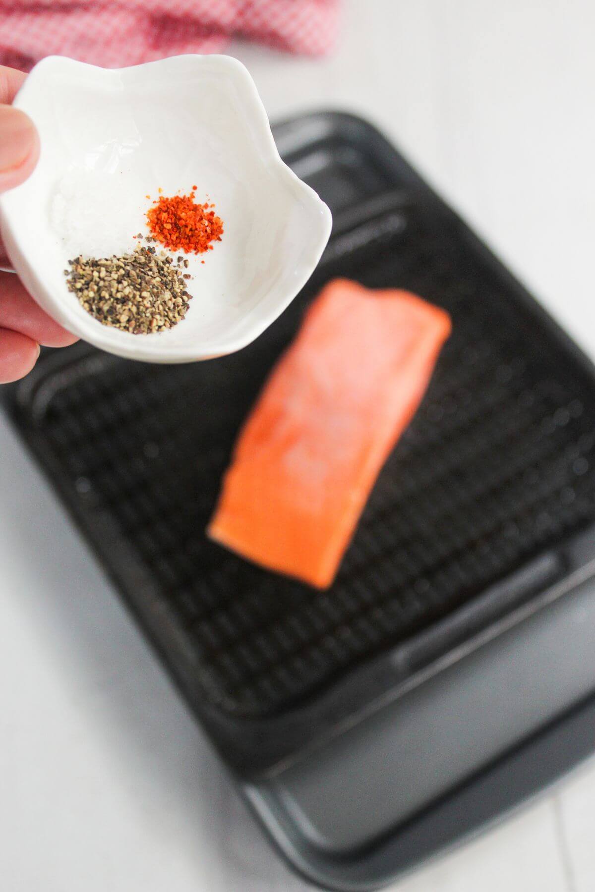 A hand holds a small dish with salt, black pepper, and red pepper over a raw salmon fillet on a grill pan.