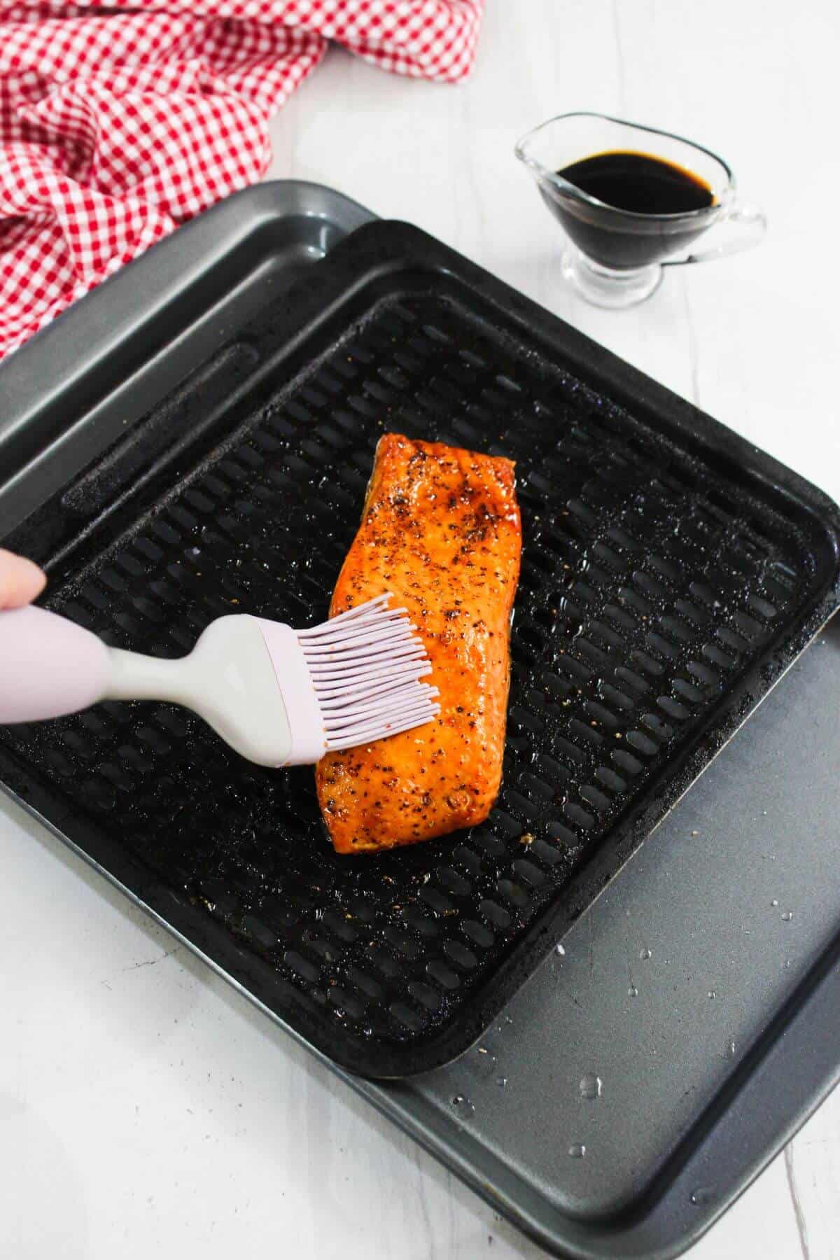 A hand brushes sauce onto a cooked salmon fillet on a grill pan, with a small pitcher of sauce and a red checkered cloth nearby.