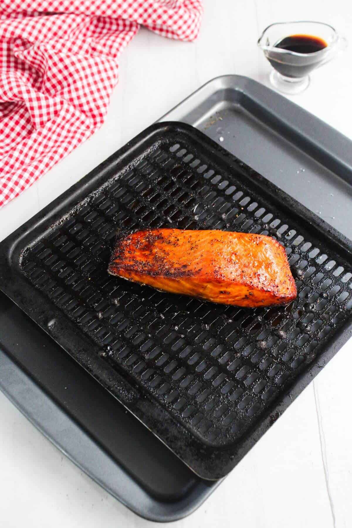 Grilled salmon fillet on a black broiler pan, resting on a baking tray, with a small glass container of sauce and a red checkered cloth nearby.