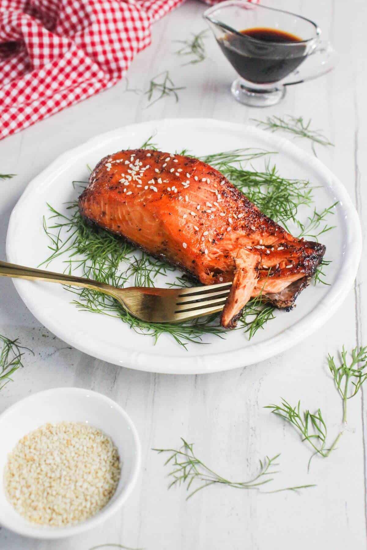 A piece of cooked salmon garnished with sesame seeds and dill on a white plate with a gold fork, a bowl of sesame seeds, a sauce boat, and a red checkered napkin nearby.