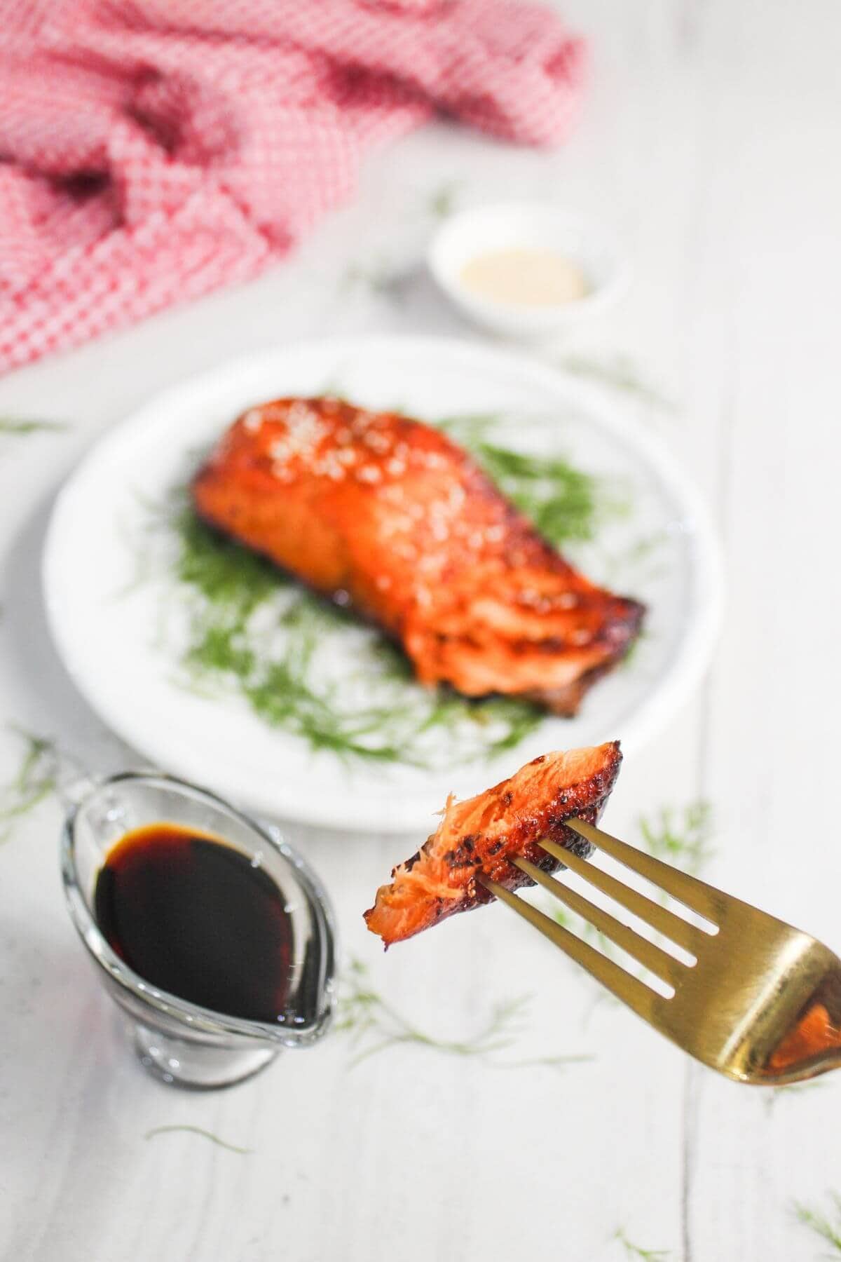 A fork holds a bite of cooked salmon above a plate with a salmon fillet on dill; a small pitcher of soy sauce and a sauce dish are in the background.