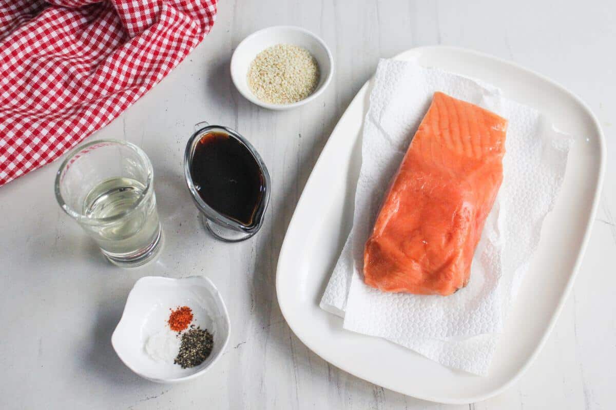 A raw salmon fillet on a plate with paper towels, next to small bowls of sesame seeds, spices, soy sauce, and a glass of clear liquid on a white surface.