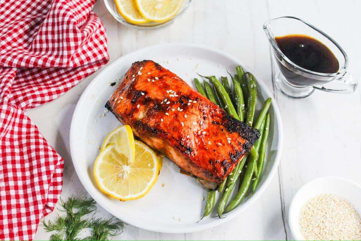 Grilled salmon fillet with sesame seeds, lemon slices, and green beans on a white plate, with sauce and a red checkered napkin beside it.