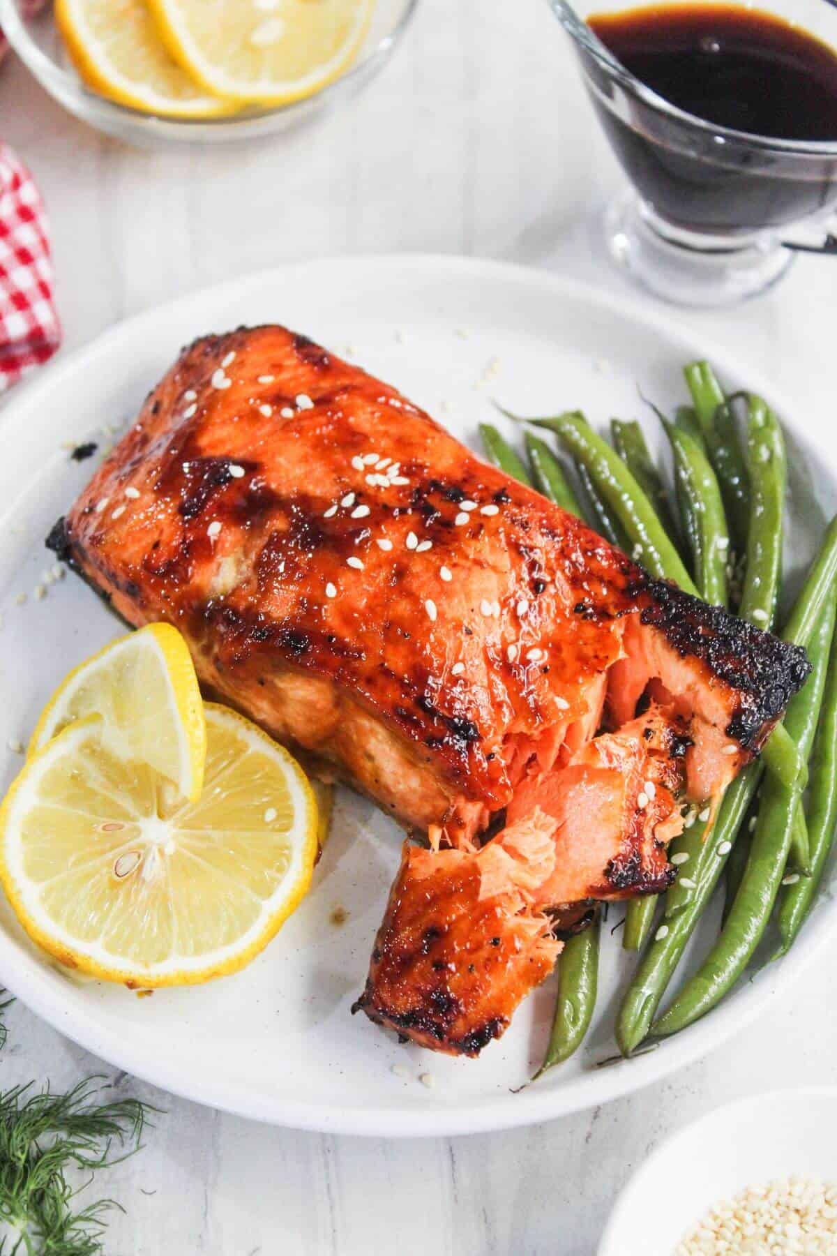 Grilled salmon fillet with a glaze, garnished with sesame seeds, served with green beans and lemon slices on a white plate.