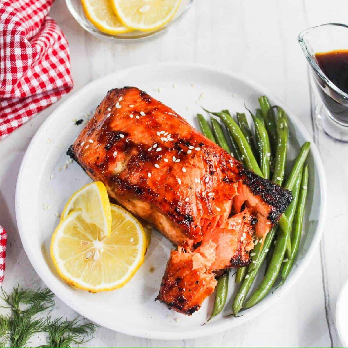 A plate with glazed salmon, green beans, and two lemon slices, served with a small pitcher of dark sauce on the side.