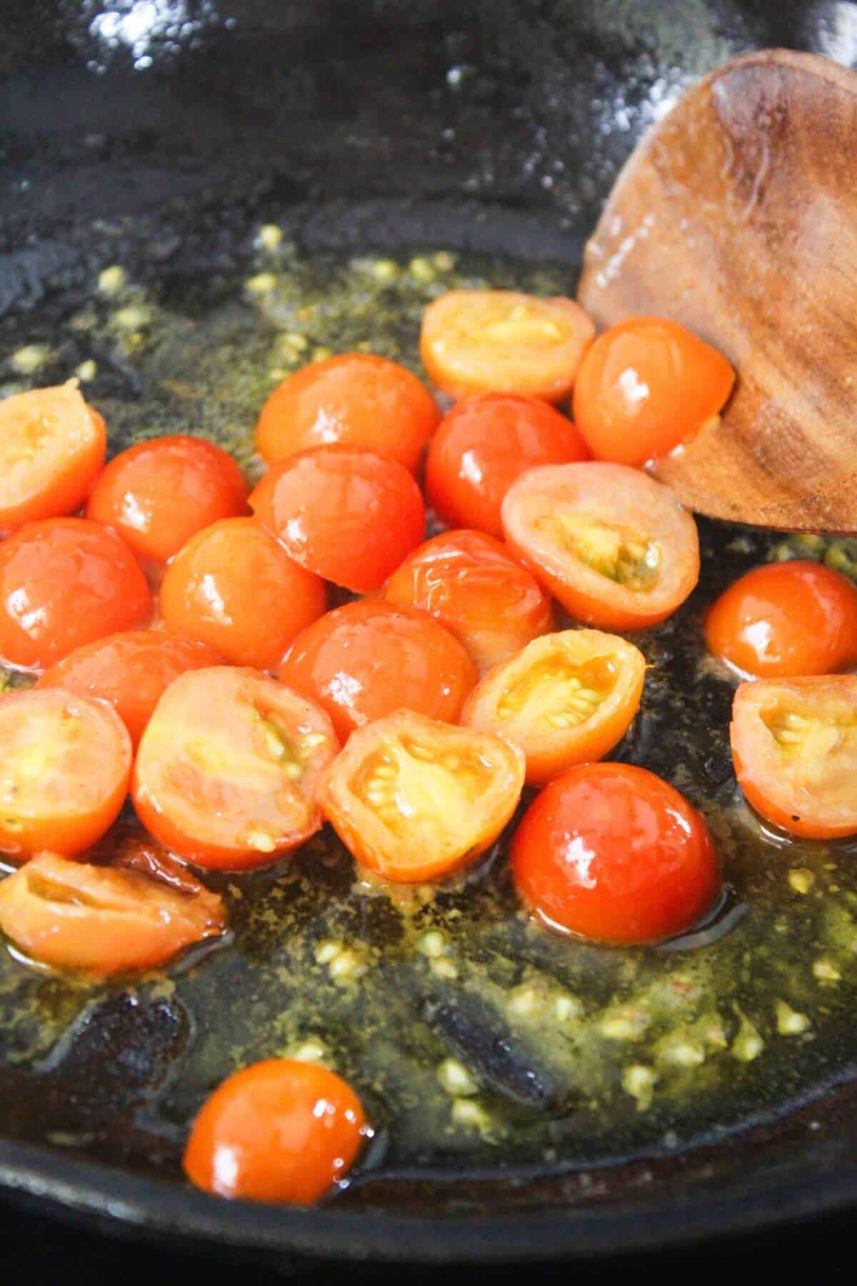 Halved cherry tomatoes cooking in a skillet with minced garlic and oil, being stirred with a wooden spoon.