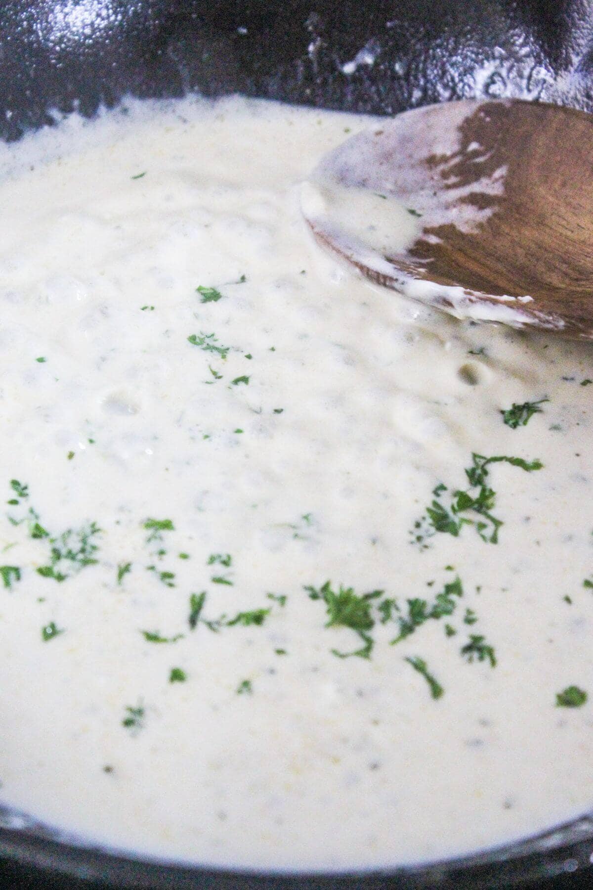 Creamy white sauce with herbs being stirred in a pan with a wooden spoon.