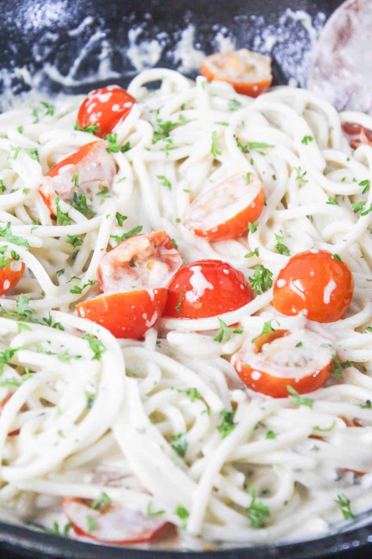 Close-up of spaghetti in a creamy white sauce with cherry tomato halves and chopped parsley, served in a pan.