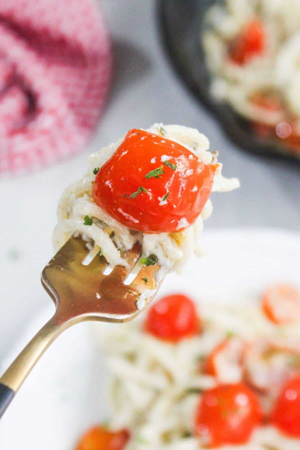 A close-up of a fork holding spaghetti and a roasted cherry tomato, with more pasta and tomatoes visible on a plate in the background.