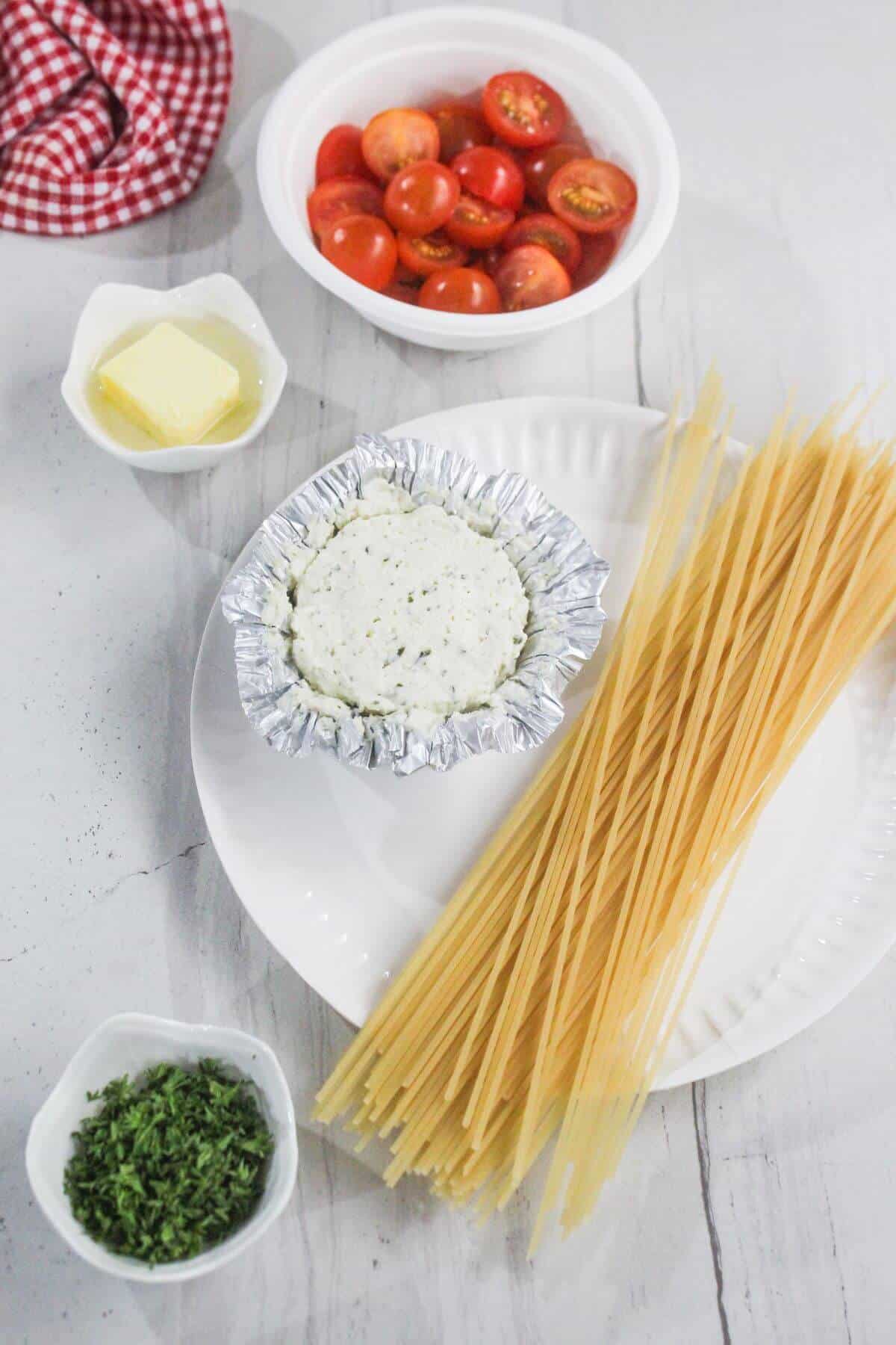 Uncooked spaghetti, a foil-wrapped cheese round, halved cherry tomatoes, butter, and chopped herbs are arranged on a white plate and table.