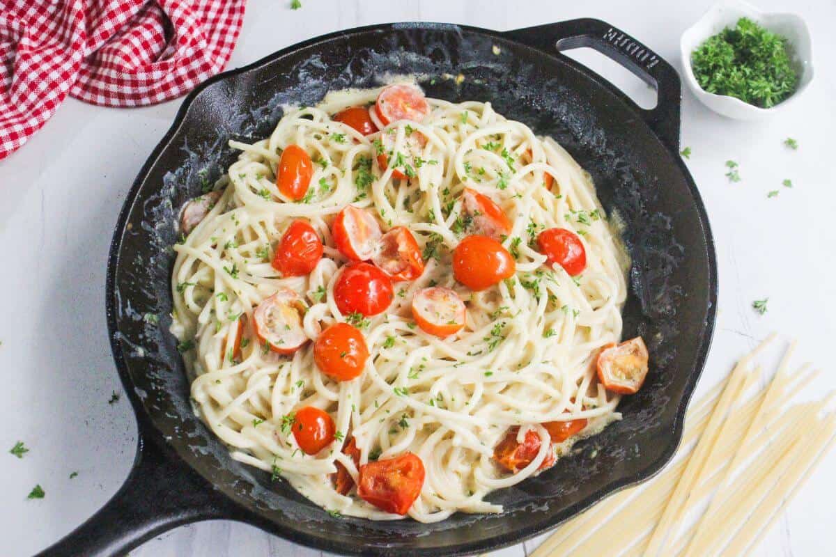A skillet filled with creamy spaghetti topped with halved cherry tomatoes and chopped herbs, with uncooked pasta and parsley nearby on a white surface.