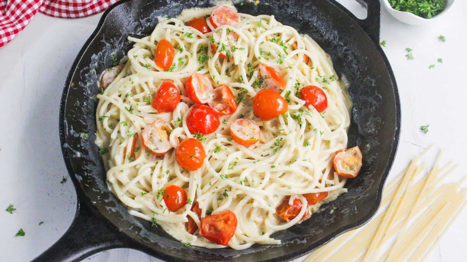 A skillet filled with creamy spaghetti, halved cherry tomatoes, and chopped herbs, with uncooked pasta and parsley visible nearby.