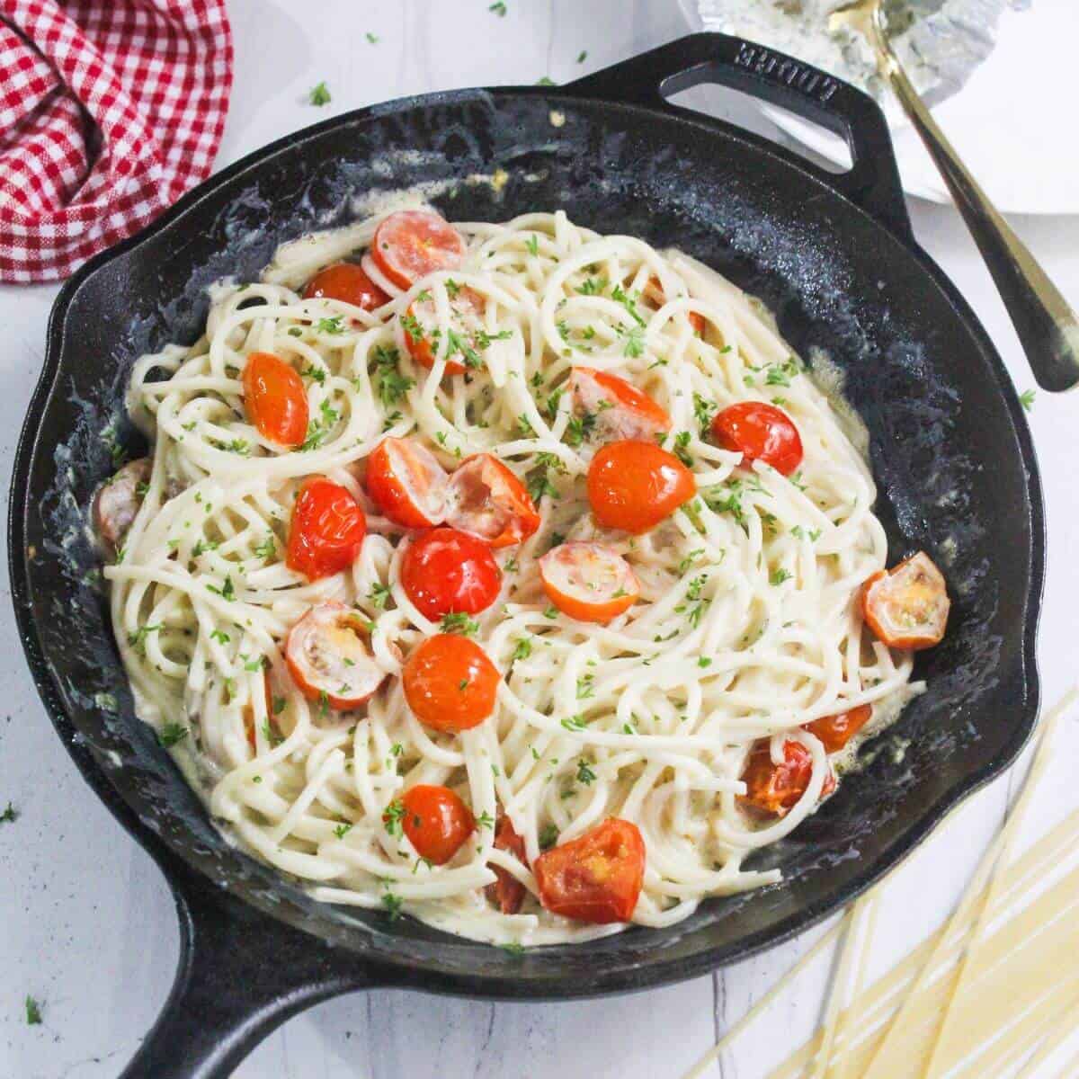 A cast iron skillet filled with spaghetti in a creamy white sauce, garnished with cherry tomatoes and chopped herbs. A red checkered cloth and utensils are nearby.