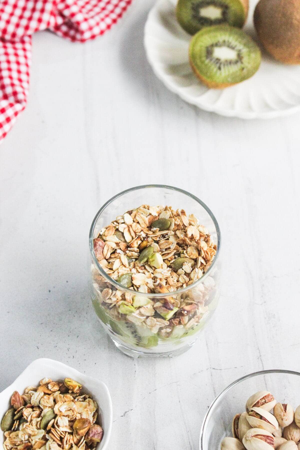 A glass filled with granola, seeds, and sliced kiwi sits on a white surface, with whole and sliced kiwis on a plate in the background.