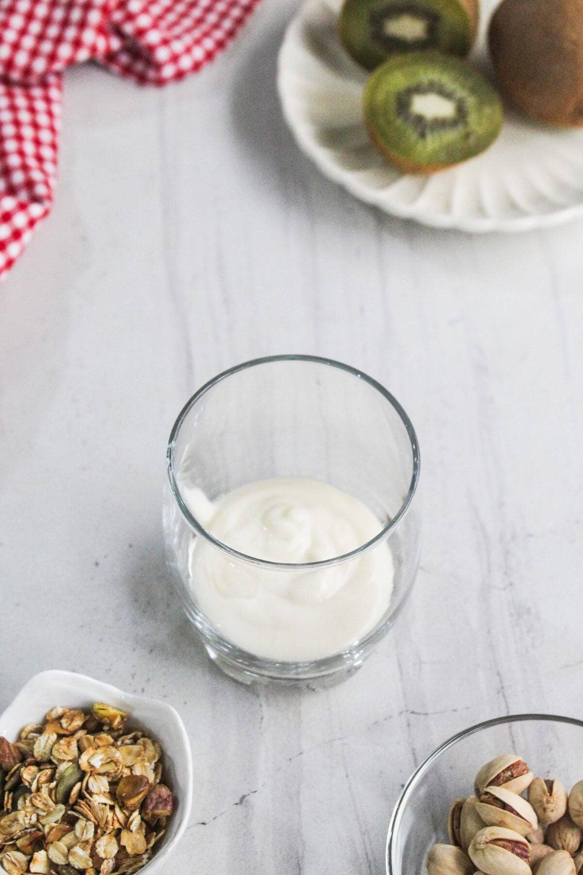 A glass with plain yogurt sits on a white surface, surrounded by a bowl of granola, pistachios, a plate with sliced and whole kiwi, and a red checkered cloth.